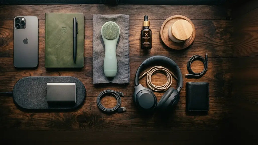 A styled flat-lay of various personal care and tech accessories on a dark wooden surface, including over-ear headphones, a smartphone, a notebook, a cordless charging pad, a small bottle, and other accessories.