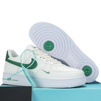 A pair of white low-top sneakers with green accents, including a green Swoosh on the side, a green heel tab, and a light blue tie detail, displayed on a light blue box with a black block. The outsole of one shoe is shown propped up behind the other.